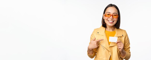 Foto grátis retrato de uma linda garota asiática moderna em óculos de sol sorrindo feliz mostrando cartão de crédito sobre o espaço de cópia de fundo branco