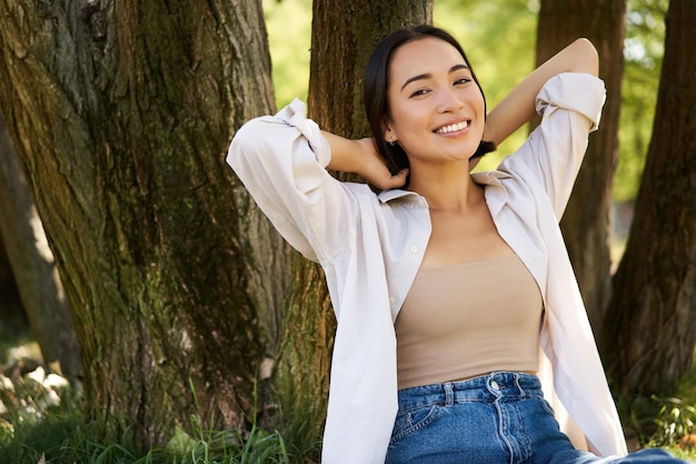 Foto grátis retrato de uma linda mulher asiática descansando perto de uma árvore relaxando no parque sorrindo e parecendo feliz