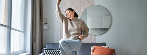 Foto grátis retrato de uma mulher asiática feliz dançando, regozijando-se e triunfando, sentindo-se otimista em casa