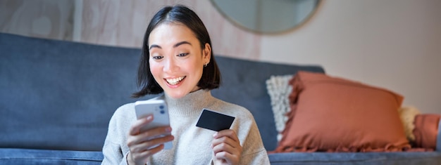 Foto grátis retrato de uma mulher asiática sorridente pagando com cartão de crédito em seu aplicativo de smartphone organiza débito direto