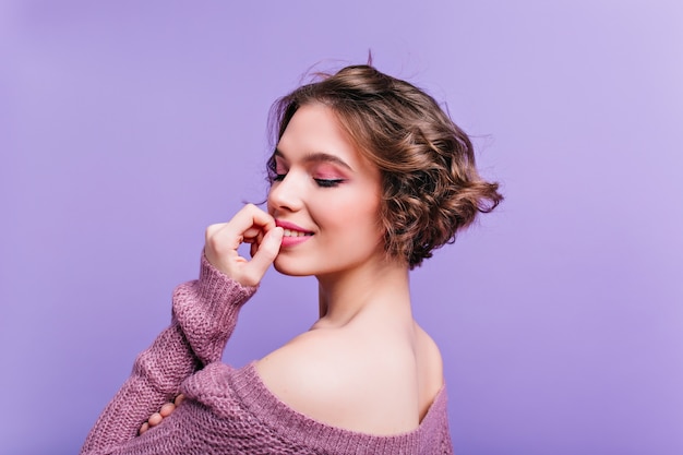 Foto grátis retrato do close-up da parte de trás do modelo feminino branco em êxtase, posando na parede roxa. menina de cabelo curto satisfeita em um traje de tricô aproveitando a sessão de fotos