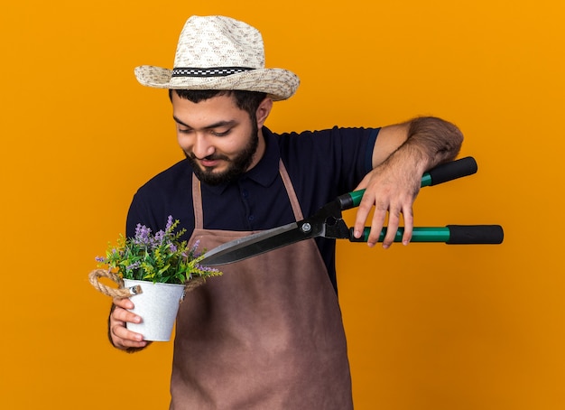 Foto grátis satisfeito jovem jardineiro caucasiano com chapéu de jardinagem segurando uma tesoura de jardinagem sobre flores em um vaso de flores isolado na parede laranja com espaço de cópia