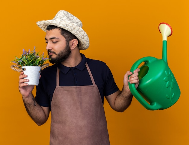 Foto grátis satisfeito jovem jardineiro caucasiano usando chapéu de jardinagem, segurando um regador e cheirando flores em um vaso de flores isolado na parede laranja com espaço de cópia