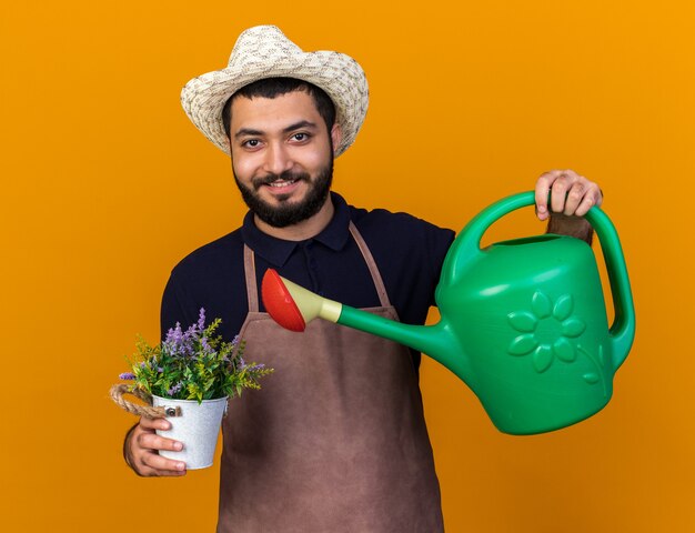 Foto grátis sorridente jovem jardineiro caucasiano com chapéu de jardinagem rega as flores em um vaso de flores com um regador isolado na parede laranja com espaço de cópia