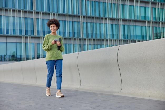 Tiro de corpo inteiro de linda mulher de cabelos cacheados usa jeans verde e tênis passeios com gadgets modernos do lado de fora perto do centro de negócios urbano tem expressão feliz Conceito de estilo de vida moderno