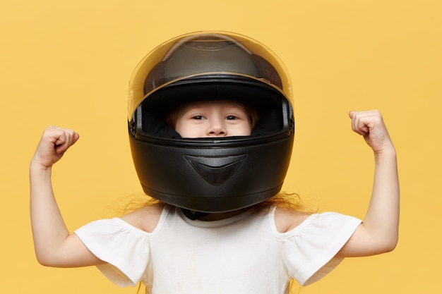 Foto grátis tiro isolado do piloto de menina posando contra uma parede amarela usando capacete preto de segurança para motocicleta, demonstrando seus músculos bíceps. conceito de pessoas, esportes radicais e adrenalina