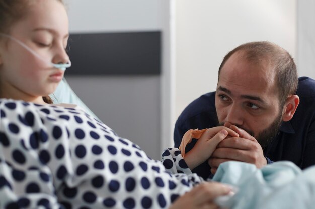 Triste e ansioso pai segurando a mão de crianças doentes dormindo na enfermaria de pacientes do hospital pediátrico. Menina doente usando tubo nasal na cama de hospital enquanto pai atento olhando para ela.