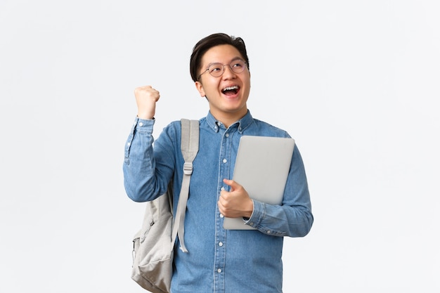 Foto grátis universidade, estudo no exterior e conceito de estilo de vida. feliz regozijando o estudante masculino asiático com aparelho triunfante, passar nos exames, terminar o semestre final, levantar o punho e gritar sim com satisfação, segurar o laptop.