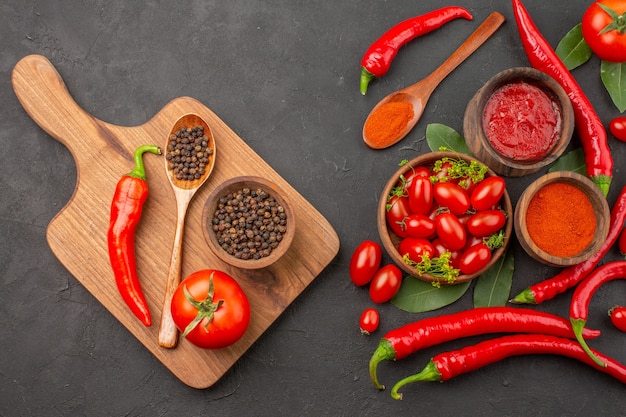 Foto grátis vista de cima uma tigela de tomate cereja, pimentão vermelho, folhas de louro e uma tigela de pimenta preta, uma colher, um tomate, uma pimenta vermelha na tábua de cortar no chão preto