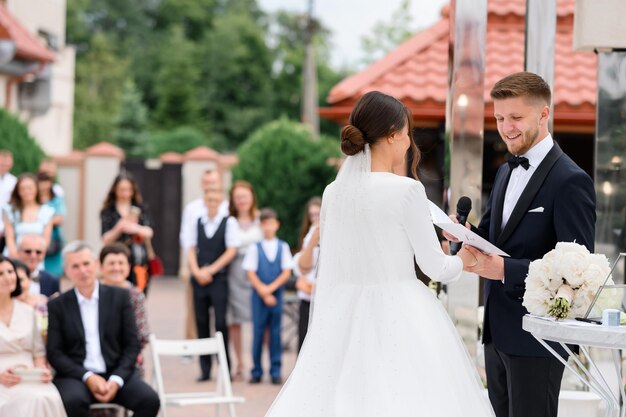 Vista lateral sorridente noivo homem com microfone segurando a mão da noiva jura um juramento na cerimônia de casamento ao ar livre Convidados felizes no fundo apreciando o lindo casal Esposa elegante no vestido inchado