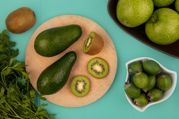 Foto grátis vista superior de abacates com fatias de kiwi em uma placa de cozinha de madeira com feijoas em uma tigela com maçãs em uma tigela em uma parede azul