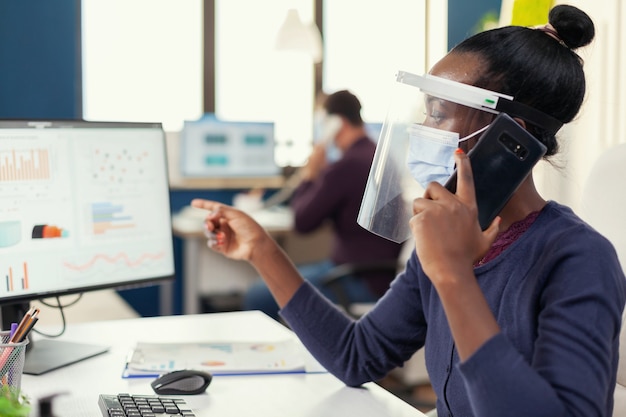 Kostenloses Foto afrikanischer mitarbeiter, der im büro auf dem smartphone spricht und eine gesichtsmaske gegen coronavirus trägt