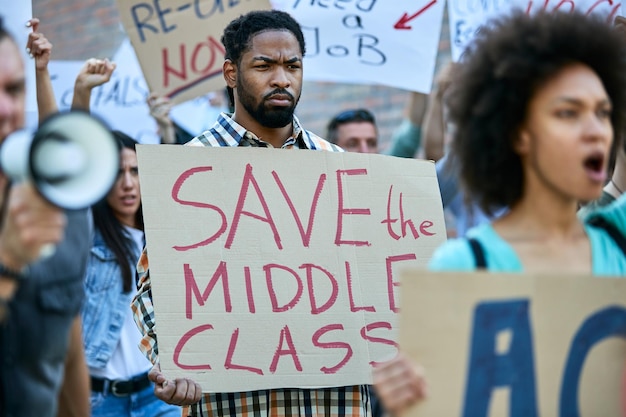 Afroamerikaner, der ein Banner mit der Aufschrift "Save the Middle Class" trägt, während er mit einer Menschenmenge auf den Straßen der Stadt protestiert