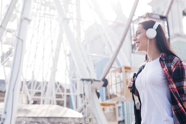 Kostenloses Foto attraktive hörende musik der jungen frau mit dem kopfhörer, der nahes riesenrad steht