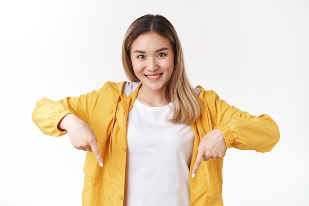 Kostenloses Foto attraktives fröhliches dummes blondes asiatisches mädchen, das nach unten zeigt zeigefinger-look-kamera, glückliches optimistisches lächeln, schlägt eine gute empfehlung vor, die weiße wand steht