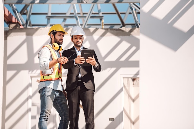 Bauingenieur und Bauarbeiter-Manager mit digitalem Tablet und Blaupausen, die über das Konzept der Zusammenarbeit auf Baustellen sprechen und planen