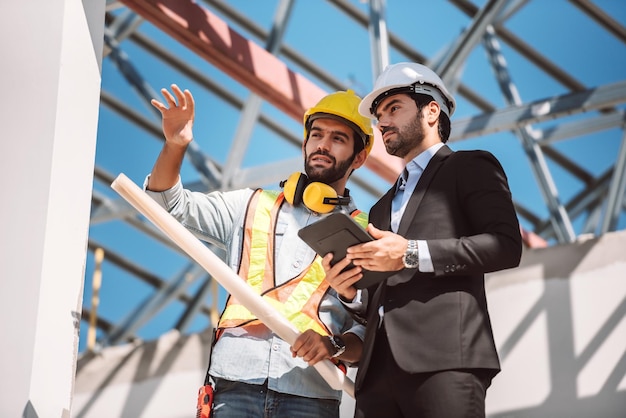 Bauingenieur und Bauarbeiter-Manager mit digitalem Tablet und Blaupausen, die über das Konzept der Zusammenarbeit auf Baustellen sprechen und planen