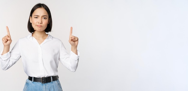 Kostenloses Foto bild einer skeptischen asiatischen dame, die verärgert die stirn runzelt und mit dem finger nach oben zeigt und werbung zeigt