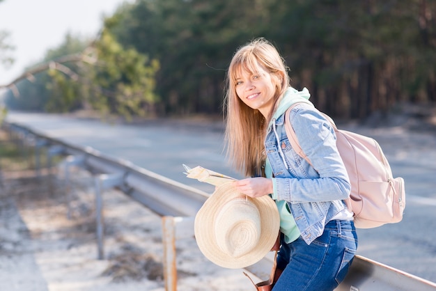 Kostenloses Foto blonde junge frau, die den hut und karte stehen nahe der straße mit ihrem rucksack betrachtet kamera hält