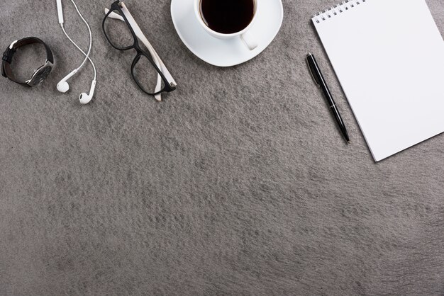 Büro grauer Schreibtisch mit Kopfhörer; Brille; Armbanduhr; Kaffeetasse; Stift und leerer Spiralblock