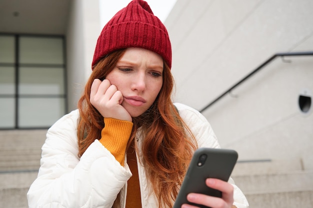 Kostenloses Foto das porträt eines traurigen rothaarigen mädchens sieht auf dem smartphone-bildschirm verärgert und enttäuscht aus und liest den text der schlechten nachricht m