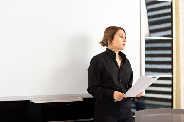 Eine junge attraktive Geschäftsfrau der Vorderansicht im schwarzen Hemd, die ihre Arbeit darstellt, die Dokumentjobaufbaupräsentation liest