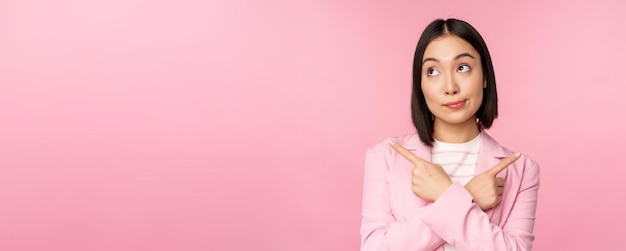 Kostenloses Foto entscheidungskonzept junge asiatische geschäftsfrau corporate worker zeigt mit dem finger seitlich in zwei richtungen und zeigt varianten, die bei der auswahl des rosafarbenen hintergrunds unentschlossen aussehen