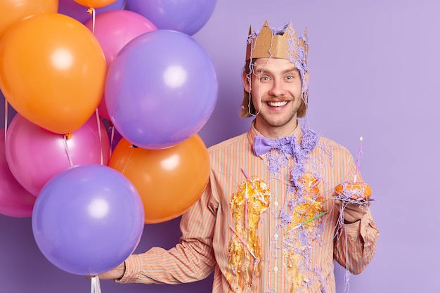 Kostenlose Foto erfreuter erwachsener mann schmutzig mit kuchencreme hält kleinen cupcake mit kerze hat spaß bei junggesellenabschied trägt papierkrone auf dem kopf hält bunte aufgeblasene luftballons isoliert über lila wand