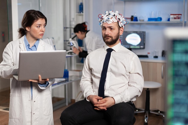 Kostenloses Foto forscherin, ärztin, die neurologische krankheitssymptome auf dem laptop eingibt und die medizinische behandlung mit dem patienten bespricht. neurologe-ingenieur, der die aktivität des nervensystems des menschen mit eeg-kopfhörer überwacht