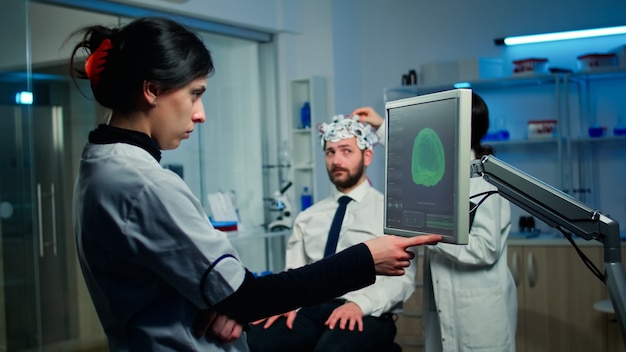 Kostenloses Foto forscherin, die auf den monitor schaut, der den gehirnscan analysiert, während die kollegin mit dem patienten im hintergrund über nebenwirkungen diskutiert