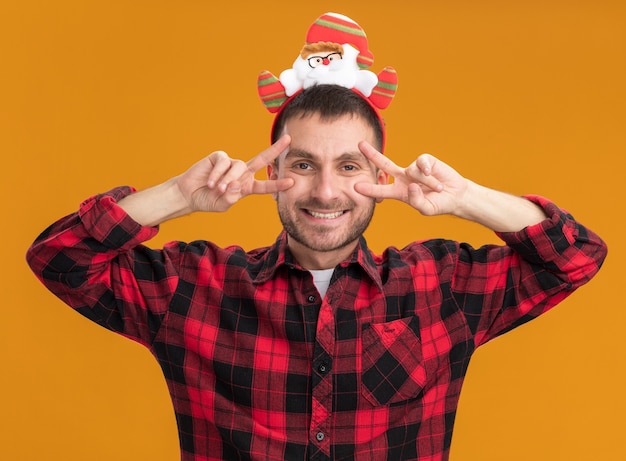 Kostenloses Foto freudiger junger kaukasischer mann, der weihnachtsmann-stirnband trägt, das v-zeichensymbole nahe den augen lokalisiert auf orange wand zeigt