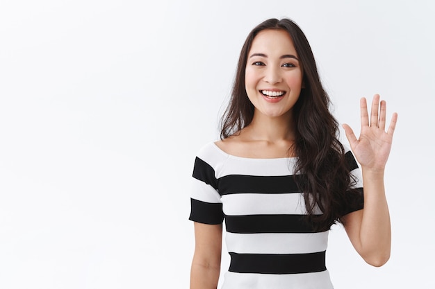 Kostenloses Foto freundliche, angenehme und fröhliche ostasiatische frau in gestreiftem t-shirt, die handfläche hebt, handgruß winkt, hallo oder hallo sagt und mit freudigem ausdruck als willkommensneulinge lächelt, weißer hintergrund