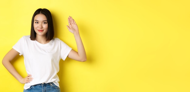 Kostenloses Foto freundliche asiatische frau in weißem t-shirt, die mit der hand winkt und hallo sagt, grüßt sie über gelb