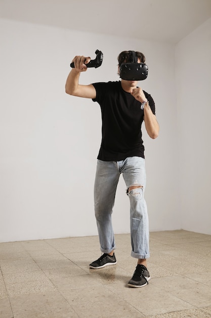 Kostenloses Foto ganzkörperporträt eines jungen spielers in vr-brille und jeans und schwarzem unbeschriftetem t-shirt, das ein spiel in einem raum mit weißen wänden und hellem holzboden spielt.