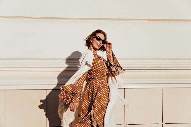 Kostenloses Foto hübsche frau im langen braunen kleid, das sonnigen tag genießt. liebenswertes weißes mädchen in der retro-kleidung, die die straße entlang geht.