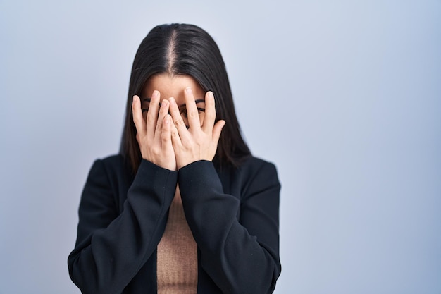 Kostenloses Foto junge brünette frau, die über blauem hintergrund steht, mit traurigem ausdruck, der das gesicht mit den händen bedeckt, während sie ein depressionskonzept weint