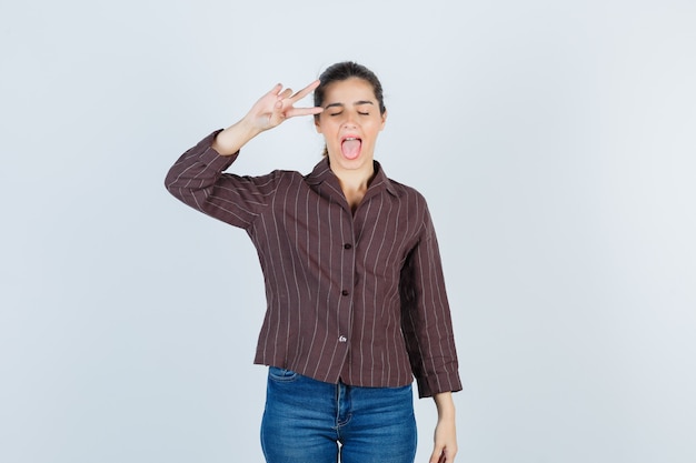 Kostenloses Foto junge schöne frau, die v-zeichen zeigt, die zunge in der jacke herausstreckt und verspielt aussieht, vorderansicht.