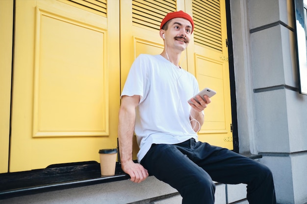 Kostenloses Foto junger attraktiver mann mit schnurrbart in t-shirt und rotem hut, der musik auf dem handy hört, während er mit gelbem hintergrund auf der stadtstraße glücklich zur seite schaut