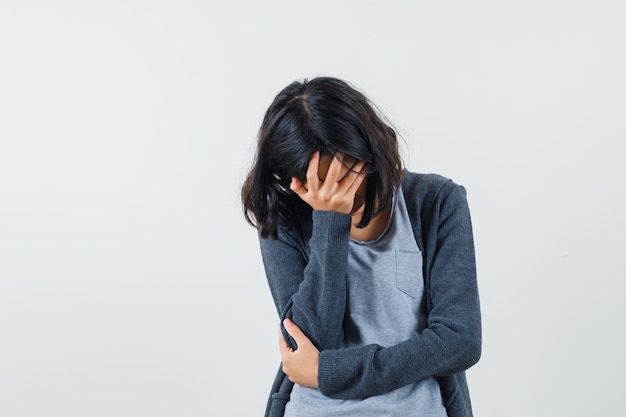 Kostenloses Foto junges mädchen, das gesicht mit hand in hellgrauem t-shirt und dunkelgrauem kapuzenpullover mit reißverschluss vorne bedeckt und gestresst aussieht.