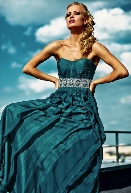 Kostenloses Foto junges sexy blondes frauenmodell im abendkleid, das auf blauem himmel aufwirft