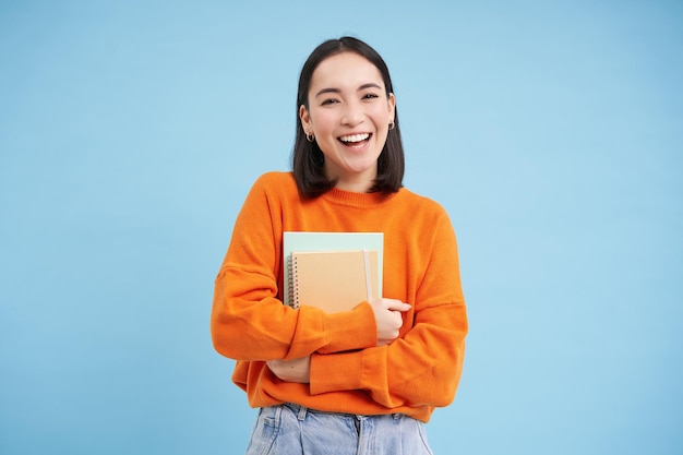 Kostenloses Foto lächelnde asiatische frau mit notizbuchstudentin mit glücklichem gesicht promo der hochschulbildung blauer hintergrund