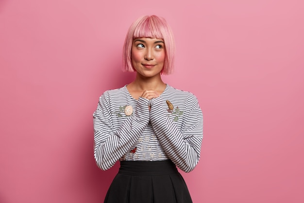 Nachdenkliche verträumte Frau mit rosa Frisur, hält die Hände zusammen, sieht geheimnisvoll aus, trägt einen modischen gestreiften Pullover und einen schwarzen Rock