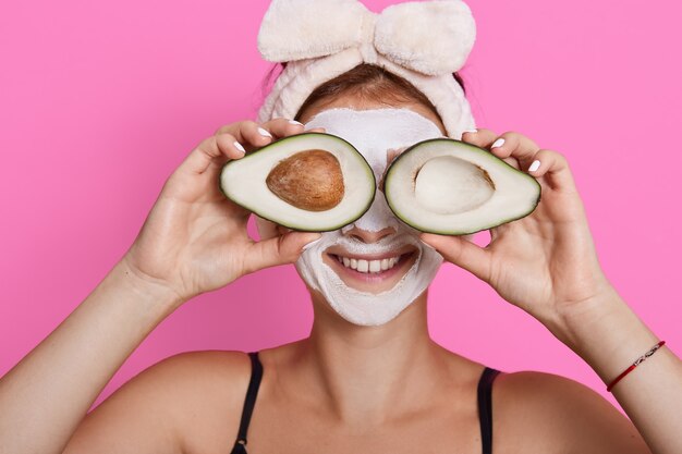 Nahaufnahmeporträt der Frau 20 mit perfekter Haut, die Avocado gegen ihre Augen lokalisiert über rosa Hintergrund, Gesundheitswesen, kosmetische Verfahren zu Hause isoliert.