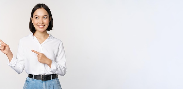 Kostenloses Foto porträt einer attraktiven erwachsenen asiatischen frau, die nach links schaut, mit einem zufriedenen lächeln, das banner oder logo beiseite zeigt, das vor weißem studiohintergrund steht