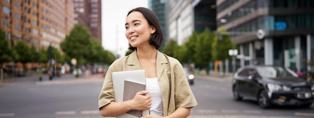 Kostenloses Foto porträt einer schönen asiatischen frau, die mit laptop und notebook auf der straße steht und zur universität geht oder