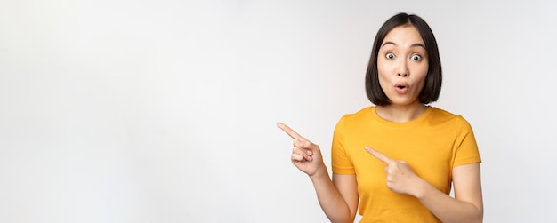 Kostenlose Foto porträt eines lächelnden asiatischen brünetten mädchens in gelbem t-shirt, das mit dem finger nach links zeigt und einen kopierraum-promo-deal zeigt, der ein banner zeigt, das über weißem hintergrund steht