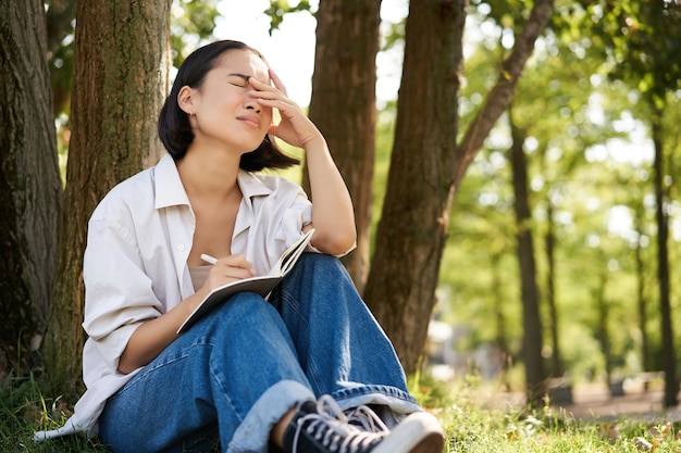 Kostenloses Foto porträt eines traurigen asiatischen mädchens, das in ihr tagebuch schreibt und sich unwohl fühlt, allein im park unter einem baum zu sitzen