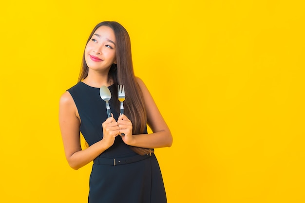 Kostenlose Foto porträt schöne junge asiatische frau bereit zu essen mit gabel und löffel auf gelbem hintergrund