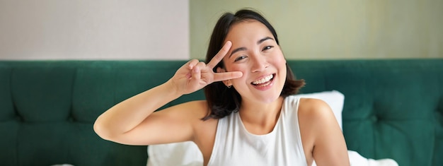 Kostenloses Foto positive asiatische frau, die im bett liegt und ein friedenszeichen zeigt, genießt einen glücklichen morgen, wenn sie optimistisch aufwacht