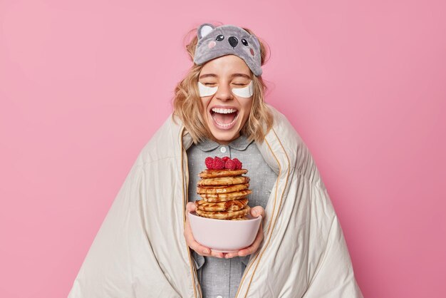 Positive optimistische Frau lacht fröhlich in Decke gehüllt hält köstliche Pfannkuchen mit Himbeere trägt eine Schlafmaske auf der Stirn trägt Schönheitsflecken unter den Augen auf, die über einer rosa Studiowand isoliert sind